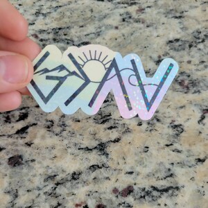 May include: A holographic sticker with a rainbow background and black outlines of a mountain, sun, and wave. The sticker says "G.O.A.T." in black letters.