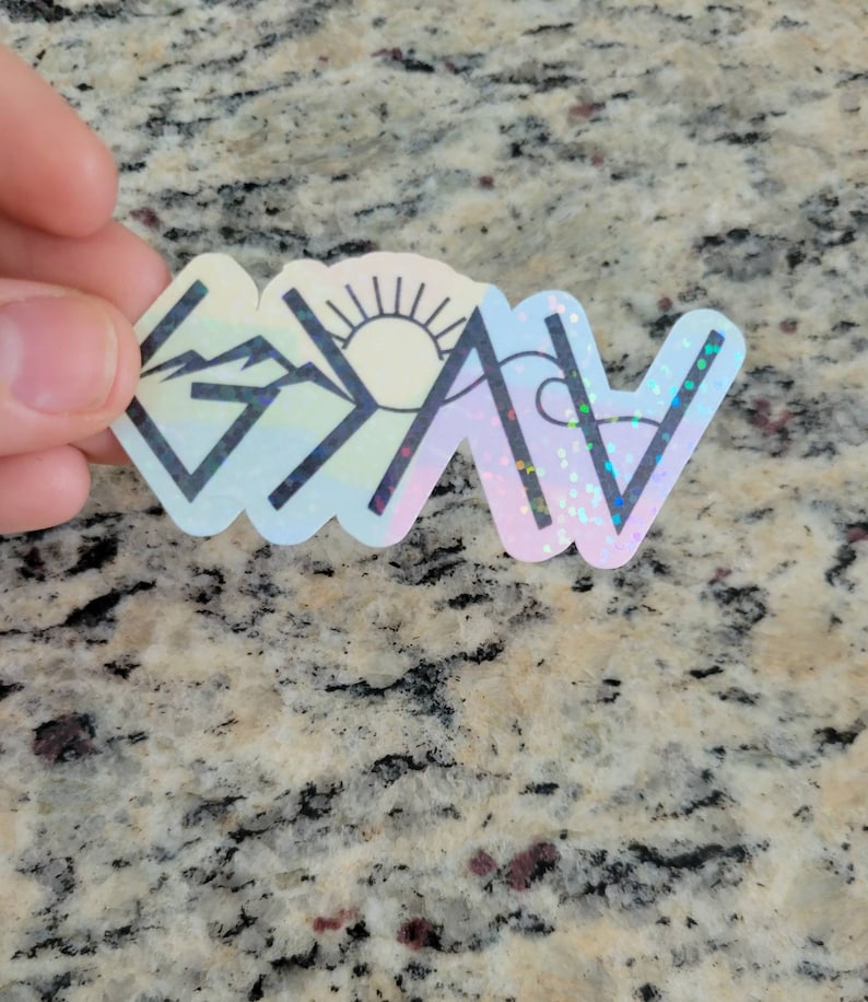 May include: A holographic sticker with a rainbow background and black outlines of a mountain, sun, and wave. The sticker says "G.O.A.T." in black letters.