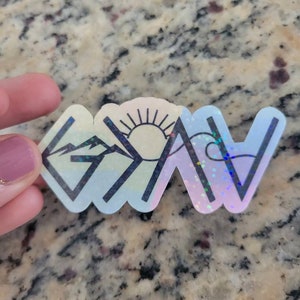 May include: A white vinyl sticker with a black outline of the word "Grateful" in a stylized font. The "G" is a mountain range, the "R" is a sun, the "A" is a wave, the "T" is a mountain range, the "E" is a wave, the "F" is a mountain range, the "U" is a sun, the "L" is a wave, and the "Y" is a mountain range. The sticker has a holographic finish.