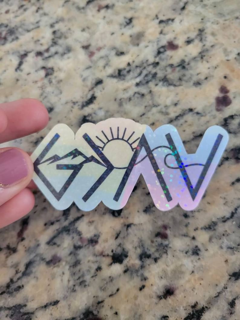 May include: A white vinyl sticker with a black outline of the word "Grateful" in a stylized font. The "G" is a mountain range, the "R" is a sun, the "A" is a wave, the "T" is a mountain range, the "E" is a wave, the "F" is a mountain range, the "U" is a sun, the "L" is a wave, and the "Y" is a mountain range. The sticker has a holographic finish.