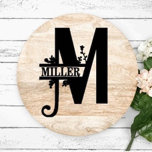 May include: A round wooden sign with a black letter "M" and the name "MILLER" in a split monogram style. The letter "M" is decorated with floral accents.