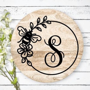 May include: A round wooden sign with a black bee and floral design, the letter "S" in a cursive font, and a black circle around the design.