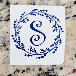 May include: A blue monogram letter "S" inside a wreath of leaves. The wreath and letter are a deep blue color, set against a white background. The design is likely a sticker or decal.