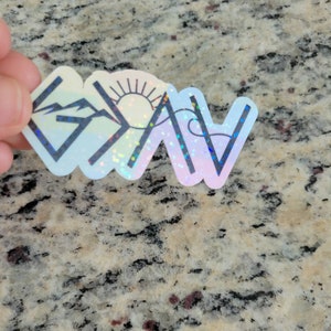 May include: A holographic sticker with a rainbow background and a black outline of the word "G.O.A.T." The "G" has a mountain range, the "O" has a sun, the "A" has a mountain range, and the "T" has a wave.