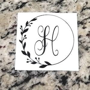 May include: A black and white sticker with a decorative wreath and the letter "H" in a cursive font.