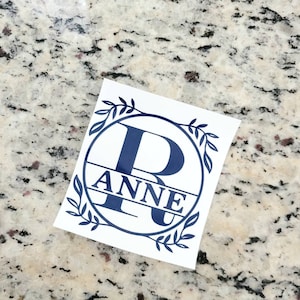 May include: A white vinyl sticker with a navy blue circle containing the letter "R" and the name "ANNE" written below the letter. The circle is surrounded by a wreath of leaves.