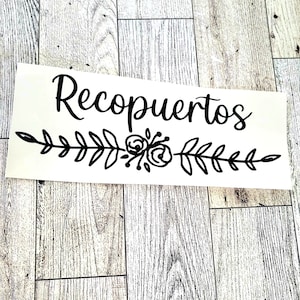 May include: Black vinyl decal with the text "Recopuertos" in a cursive font. The text is centered above a black floral design with leaves and flowers.