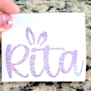 May include: A white vinyl sticker with the name "Rita" written in a cursive font with a glittery purple finish. The name is topped with a glittery purple bunny ear design.
