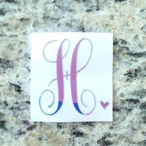 May include: A white vinyl decal featuring a large, stylized letter "H" in a gradient of pink and blue. A small pink heart is to the right of the letter. The decal is designed for application on various surfaces.
