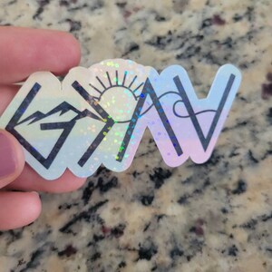 May include: A holographic sticker with a mountain, sun, and wave design. The sticker is white with a rainbow iridescent finish. The words "Grateful" are written in black.