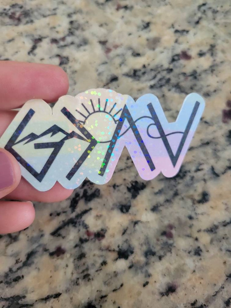 May include: A holographic sticker with a mountain, sun, and wave design. The sticker is white with a rainbow iridescent finish. The words "Grateful" are written in black.