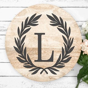 May include: A round wooden sign with a black wreath and the letter "L" in the center. The sign is on a white wooden background.