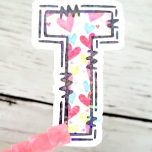 May include: A white sticker with a black outline of the letter "T". The letter is filled with a pattern of pink, blue, and yellow hearts.