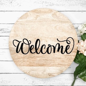 May include: A round wooden sign with the word "Welcome" written in black cursive script. The sign is on a white wooden background with white and pink flowers.