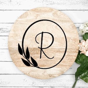 May include: A round wooden serving tray with a black circle and the letter "R" in the center. The circle is surrounded by a black leafy design.
