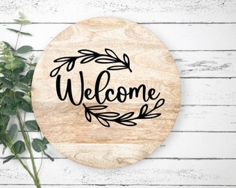 May include: A round wooden sign with the word "Welcome" written in black cursive lettering. The word is surrounded by a wreath of black leaves.