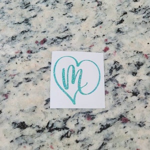 May include: A teal blue glitter heart-shaped decal with the letter "M" inside. The decal is on a white background.