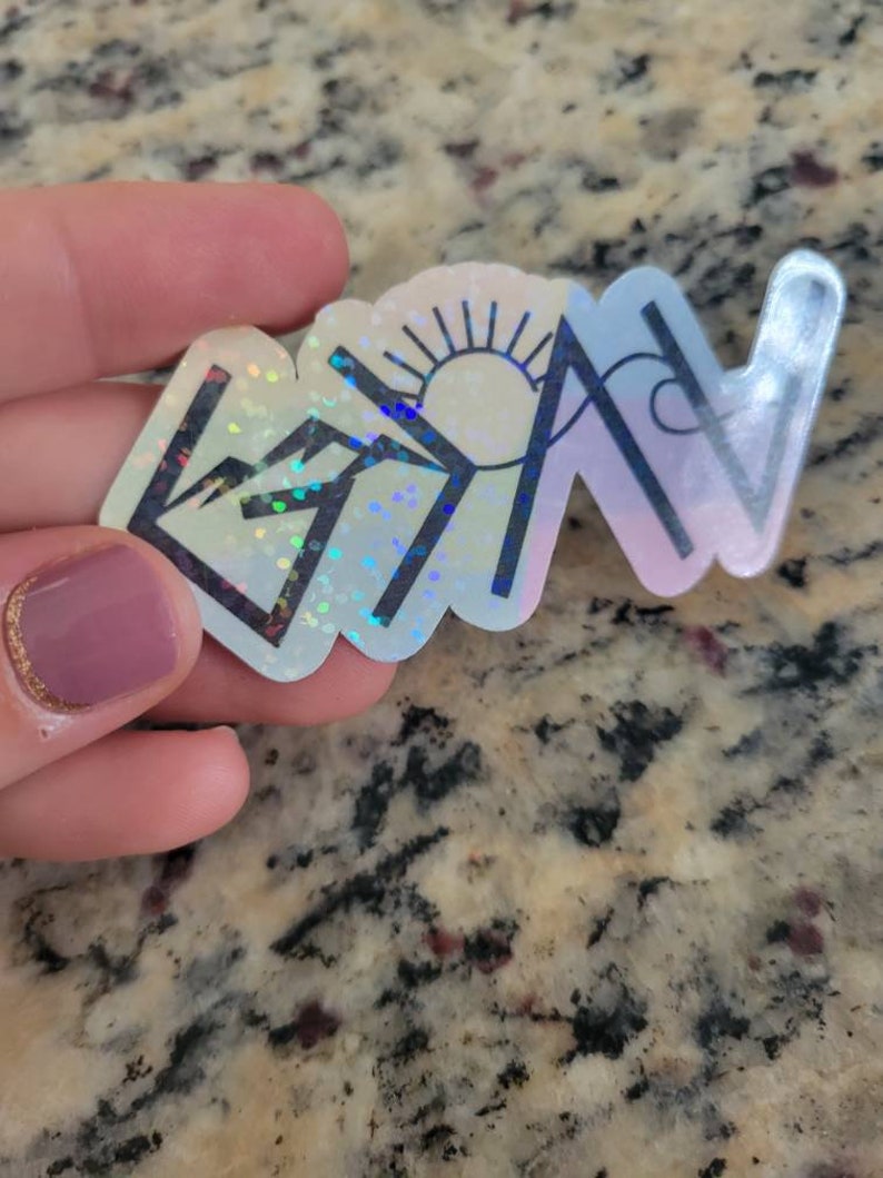 May include: A holographic sticker with a mountain, sun, and wave design. The sticker is iridescent and has a rainbow effect.
