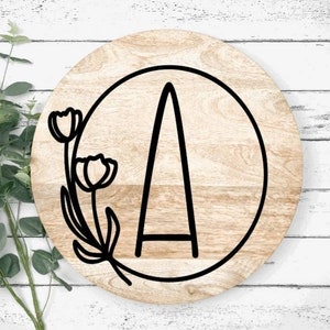 May include: A wooden round sign with a black outline circle and the letter "A" inside. The circle is decorated with a floral design with two tulips.