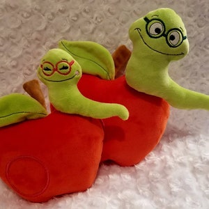 May include: Two plush green worm toys with glasses, each sitting on a red plush apple. The apples have green leaves.