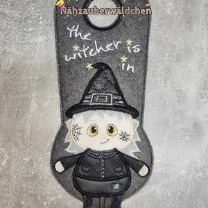 May include: A gray felt door hanger with a black and white witch illustration. The text "The witch is in" is written in white on the door hanger.