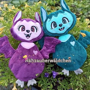 May include: Two plush bat toys, one purple and one teal, with embroidered eyes and mouths. Both have wings and feet. The purple bat has a pink heart on its chest and the teal bat has a white heart on its chest. The text "Nähzauberwäldchen" is embroidered on the purple bat's chest.