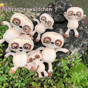 Puede incluir: Cinco peluches de perezosos de diferentes tamaños, todos con pelaje marrón, caras sonrientes y grandes ojos verdes. Los perezosos están dispuestos en una roca y en follaje verde.