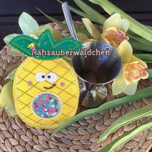 Könnte beinhalten: Eine gelbe Filzananas mit grünem Filz-Oberteil und einem lächelnden Gesicht. Die Ananas hat eine glitzernde Mitte mit bunten Konfetti. Der Text "Nähzauberwäldchen" ist auf die Ananas geschrieben.