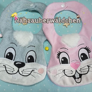 May include: Two baby bibs with bunny faces embroidered on them. The bib on the left is light blue with a white bunny face. The bib on the right is pink with a white bunny face.