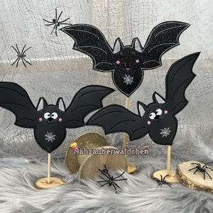 May include: Three black bat decorations with smiling faces and spiderweb designs, mounted on wooden sticks. The bats have outstretched wings and are set against a backdrop with spiderweb accents and small spider figurines. The bats are on wooden bases.