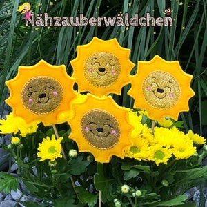 Puede incluir: Cuatro decoraciones de sol de fieltro amarillo con caras sonrientes y detalles de purpurina. Los soles están en palos y están dispuestos en una maceta con flores amarillas.