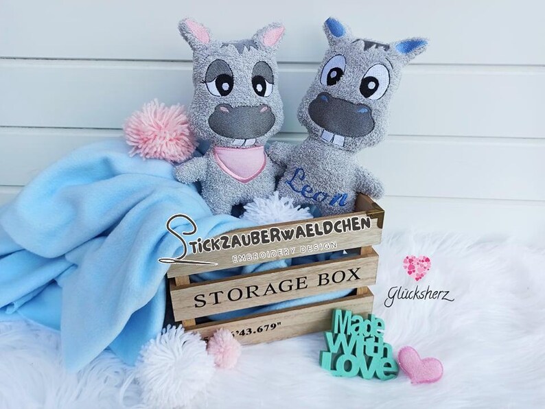K&ouml;nnte beinhalten: Zwei graue Pl&uuml;schtiere mit gestickten Gesichtern und den Namen Leon und Stickzauberwaeldchen. Die Spielzeuge sitzen in einer Holzkiste mit dem Text "Storage Box" auf der Vorderseite.