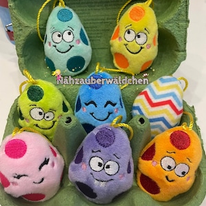 May include: Six plush Easter eggs in a green egg carton. The eggs are various colors and have different embroidered faces. The text "Nähzauberwäldchen" is visible on the carton.