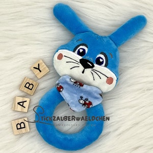 Puede incluir: Sonajero de peluche azul en forma de conejo con una bufanda de estampado azul y blanco. El sonajero tiene forma de anillo y tiene una peque&ntilde;a cara bordada con ojos negros y una nariz negra. Las palabras "BABY" est&aacute;n escritas en bloques de madera.