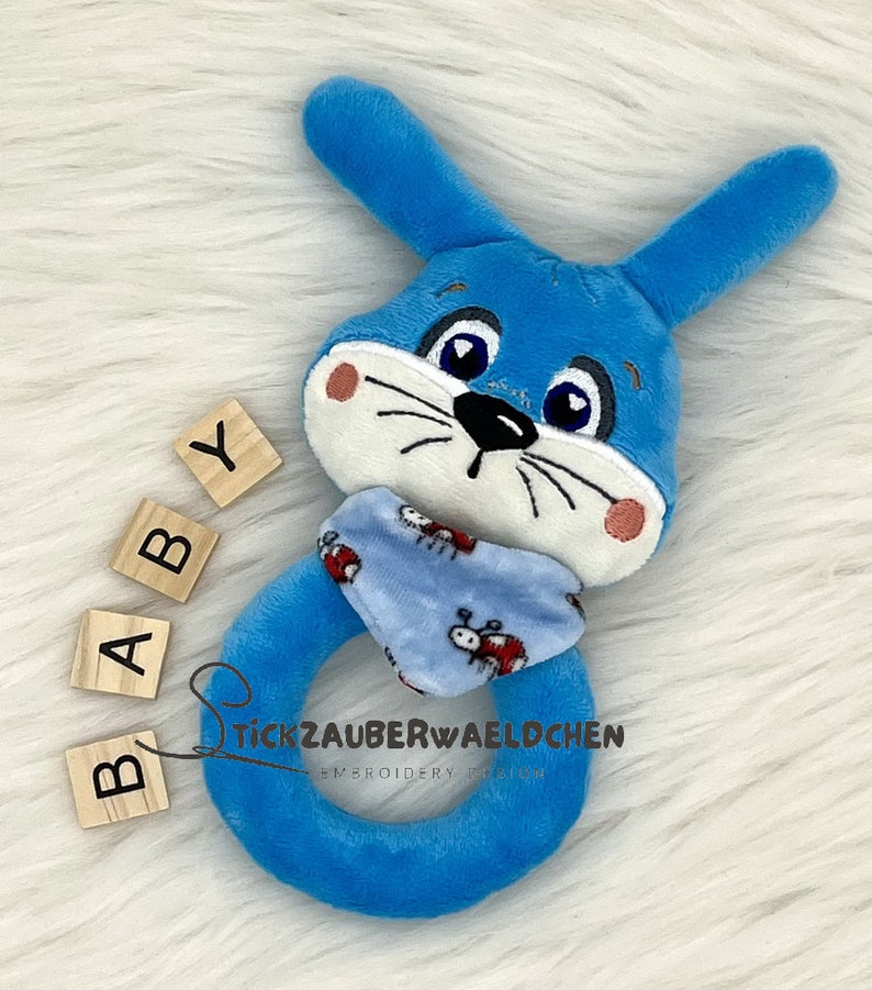Puede incluir: Sonajero de peluche azul en forma de conejo con una bufanda de estampado azul y blanco. El sonajero tiene forma de anillo y tiene una peque&ntilde;a cara bordada con ojos negros y una nariz negra. Las palabras "BABY" est&aacute;n escritas en bloques de madera.