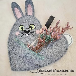 Könnte beinhalten: Ein grauer Filz-Herz-Hase mit rosa Ohren und grünen Augen. Der Hase hat eine Tasche an der Seite und hält einen Strauß getrockneter Blumen. Der Hase ist ein handgemachtes Stück und perfekt für ein Geschenk oder eine Dekoration.