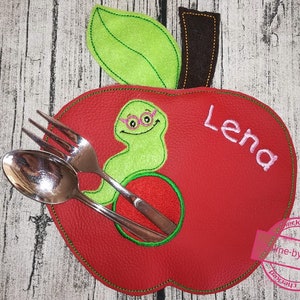 May include: Red apple-shaped placemat with a green leaf and a green worm with glasses. The placemat has the name "Lena" embroidered on it.