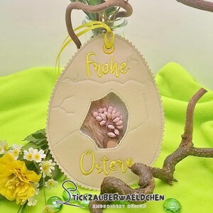 Könnte beinhalten: Ein beigefarbener Filz-Osterei-Anhänger mit gelber Stickerei, auf der "Frohe Ostern" steht, und einem kleinen Fenster mit rosa Blüten.