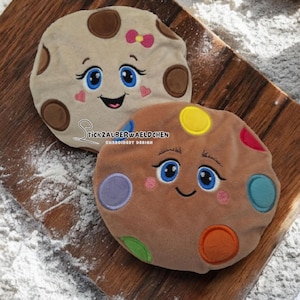 May include: Two plush cookie-shaped pillows with embroidered faces. One is tan with brown spots and a pink bow. The other is brown with colorful spots. The pillows have blue eyes, pink cheeks, and smiling mouths. The text "Stickzauberwaeldchen" is visible.