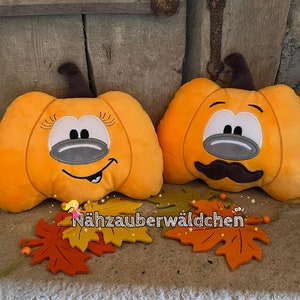 May include: Two orange plush pumpkin pillows with embroidered faces. The pumpkin on the left has a smiling face with eyelashes and the pumpkin on the right has a mustache and a serious expression. The pillows are sitting on a wooden surface with fall leaves.