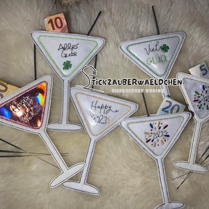 May include: Six white felt martini glass ornaments with embroidered designs. The designs include "Happy 2023", "Alles Gute", "Viel Glück", and fireworks. Each glass has a slot for a bill, and some have bills inserted. The glasses are on a white fuzzy surface.