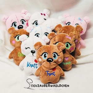 Puede incluir: Un grupo de peluches de varios colores, incluyendo rosa, blanco y marrón. Los peluches son todos osos con caras bordadas y diferentes nombres, incluyendo "Kraft", "Trust" y "Glücksbringer".