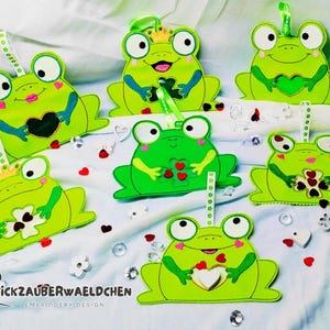 Könnte beinhalten: Sechs grüne Filz-Frosch-Anhänger mit Stickdetails. Jeder Frosch hält ein Herz oder ein Kleeblatt. Die Anhänger sind mit grünen, roten und weißen Akzenten verziert. Der Text "Stickzauberwaeldchen Embroidery Design" ist in der linken unteren Ecke sichtbar.