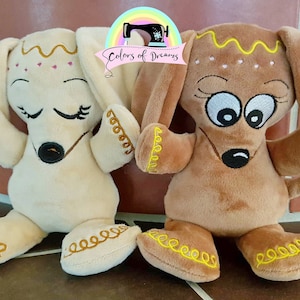 May include: Two plush dog toys, one beige and one brown, with embroidered details. Both have yellow stitching around their paws and ears. The beige dog has pink hearts and closed eyes, while the brown dog has large eyes and a frown.