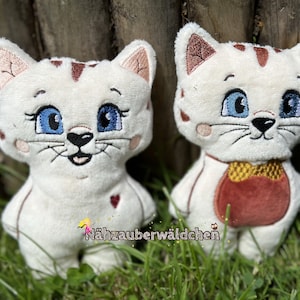 Op de afbeelding: Twee witte pluche katten speelgoed met geborduurde details. Eén kat heeft een geborduurd rood en gouden schortje, de andere heeft een rood hart op zijn borst. Beide katten hebben blauwe ogen en roze neuzen.