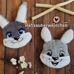 Può includere: Due teste di coniglio di peluche grigie con volti ricamati. I conigli hanno orecchie bianche e nasi neri. Il testo "Nähzauberwäldchen" è visibile sullo sfondo.