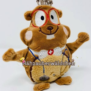 May include: A brown plush toy squirrel wearing red glasses and a blue stethoscope. The squirrel has a white embroidered smile and a white cross on its chest. The toy is labeled "Nähzauberwäldchen".