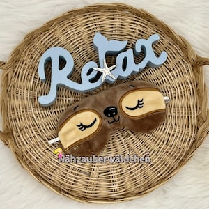 Könnte beinhalten: Ein Weidenkorb mit einem blauen Holzschild mit der Aufschrift "Relax" und einer braunen Schlafmaske mit Wimpern.