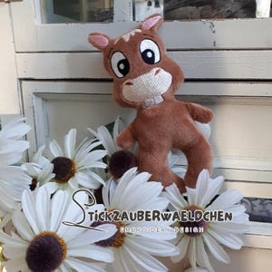 K&ouml;nnte beinhalten: Ein braunes Pl&uuml;schpferd mit wei&szlig;em Gesicht und gro&szlig;en Augen. Das Pferd steht auf einer wei&szlig;en Oberfl&auml;che mit wei&szlig;en Blumen.