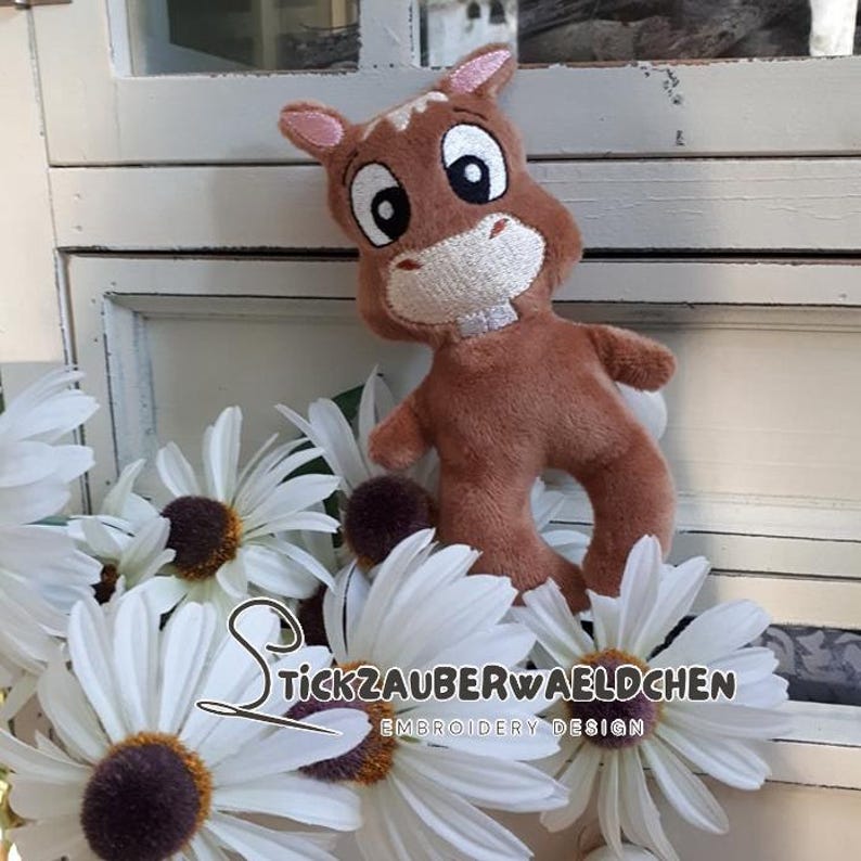 K&ouml;nnte beinhalten: Ein braunes Pl&uuml;schpferd mit wei&szlig;em Gesicht und gro&szlig;en Augen. Das Pferd steht auf einer wei&szlig;en Oberfl&auml;che mit wei&szlig;en Blumen.